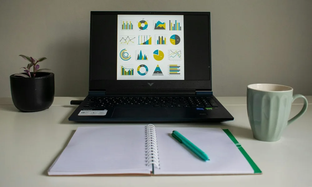 A laptop displaying analytics charts beside a notebook, green pen, and coffee mug